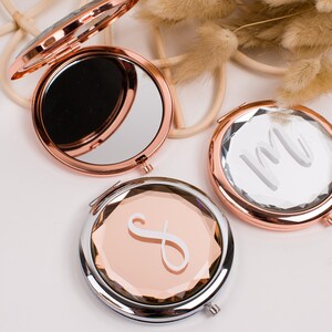 May include: Three round, rose gold compact mirrors with a faceted design. The mirrors have a silver trim and are engraved with different initials.