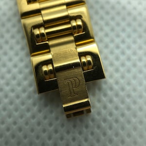 May include: Close-up of a gold watch band with a clasp that has the letter "P" engraved on it.