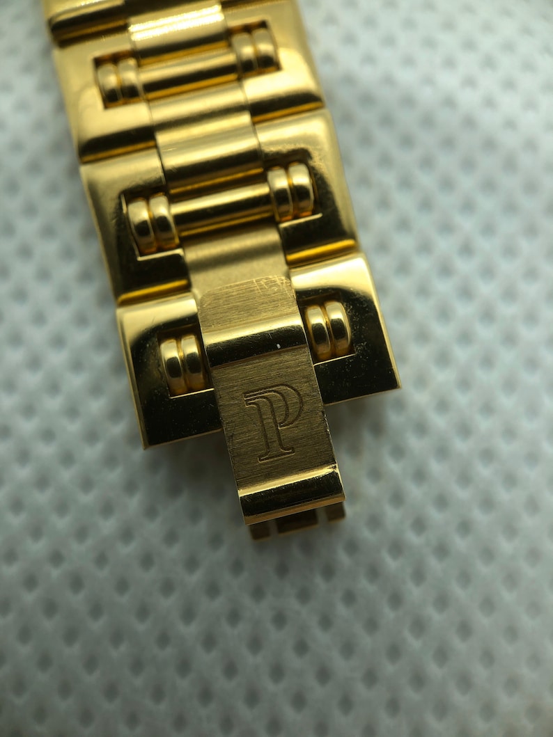 May include: Close-up of a gold watch band with a clasp that has the letter "P" engraved on it.