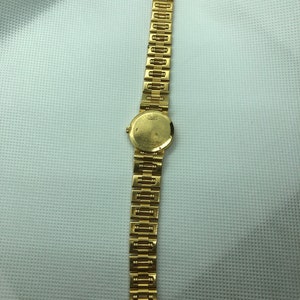 May include: A gold wristwatch with a round face and a gold metal band. The watch face has a white background and black markings. The band is made of interlocking links.