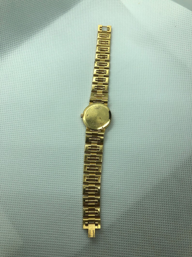 May include: A gold wristwatch with a round face and a gold metal band. The watch face has a white background and black markings. The band is made of interlocking links.