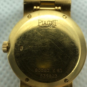 May include: Gold wristwatch with a round face and a gold band. The back of the watch has the inscription "PIAGET" and the numbers "80563 K 81 539823".