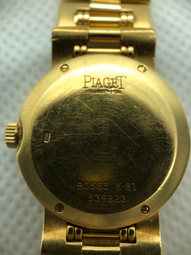 May include: Gold wristwatch with a round face and a gold band. The back of the watch has the inscription "PIAGET" and the numbers "80563 K 81 539823".
