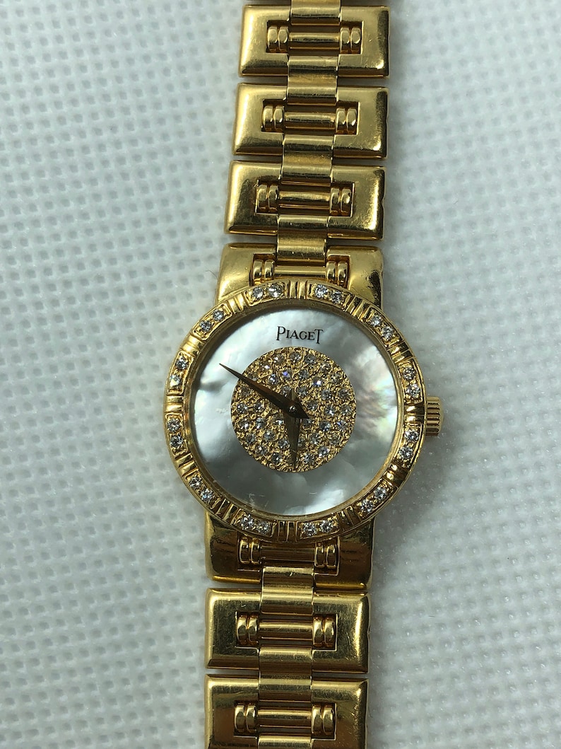 May include: A gold Piaget watch with a white mother-of-pearl face and diamond accents. The watch has a gold band.