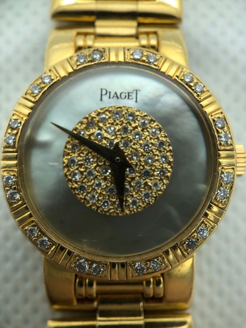 May include: A gold Piaget watch with a white mother-of-pearl face and a diamond-encrusted bezel. The watch has a gold band and a diamond-encrusted dial.