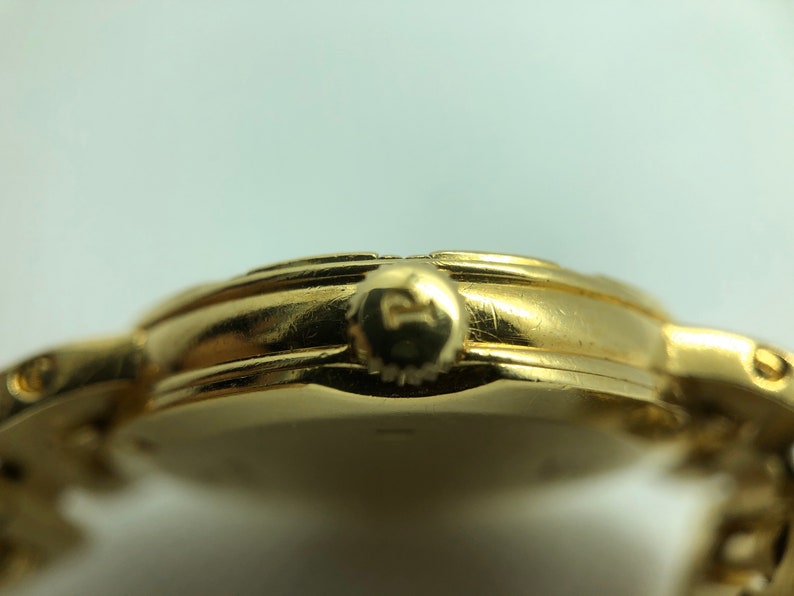 May include: A gold-toned wristwatch with a round face and a metal band. The watch has a small crown on the side.