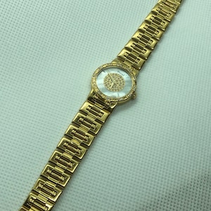 May include: A gold-tone wristwatch with a white face and a diamond-like pattern. The watch has a gold-tone band with a repeating pattern.