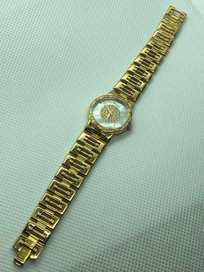 May include: A gold-tone wristwatch with a white face and a diamond-like pattern. The watch has a gold-tone band with a repeating pattern.