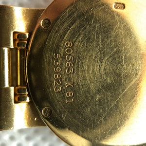 May include: Close-up of the back of a gold-tone wristwatch with the inscription "18 K 80563 53282" engraved on the case.