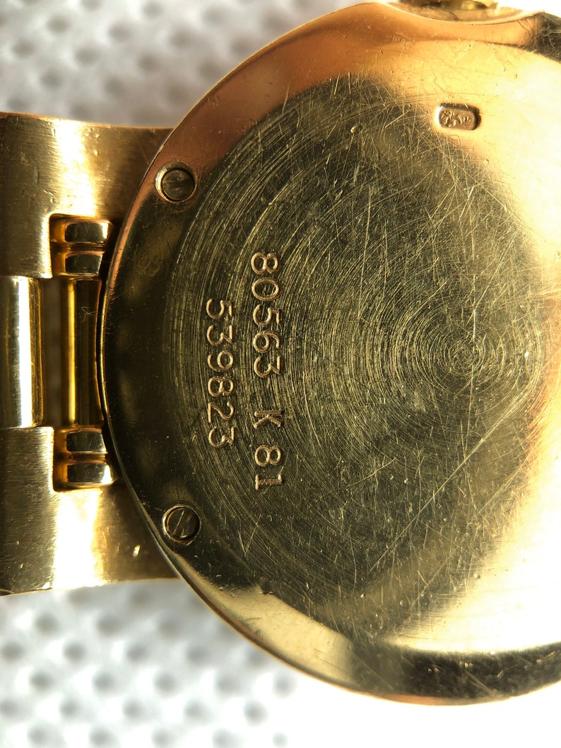 May include: Close-up of the back of a gold-tone wristwatch with the inscription "18 K 80563 53282" engraved on the case.