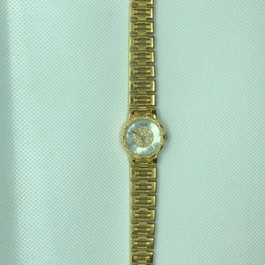 May include: A gold-tone wristwatch with a round, white face and a gold-tone band. The face is decorated with small, clear stones.