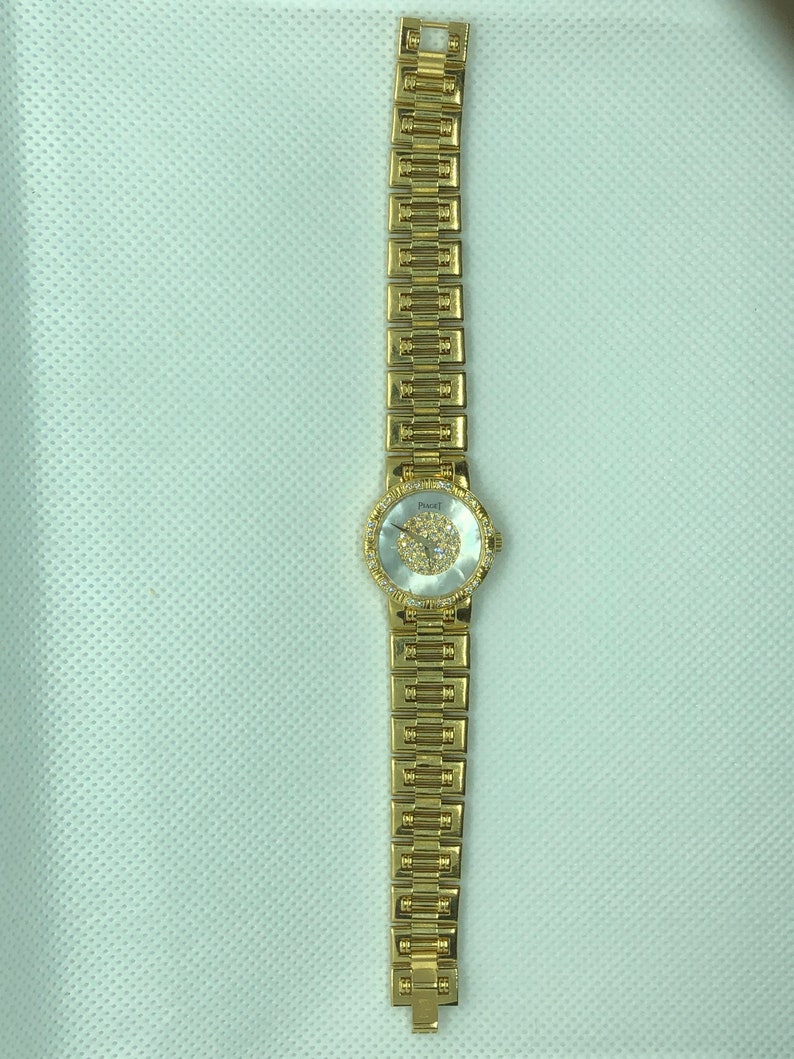 May include: A gold-tone wristwatch with a round, white face and a gold-tone band. The face is decorated with small, clear stones.