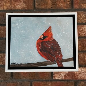 Op de afbeelding: Een ingelijst schilderij van een rode kardinaal die op een tak zit tegen een besneeuwde achtergrond. Het schilderij is onderaan links gesigneerd met "M.K.K.Liz".