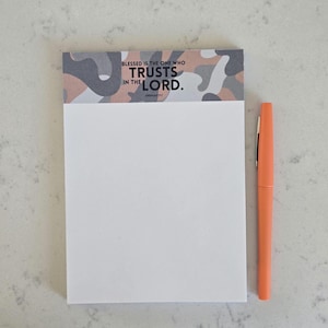 May include: A white notepad with a camouflage pattern at the top, featuring the text "BLESSED IS THE ONE WHO TRUSTS IN THE LORD." An orange pen rests beside the notepad. The notepad and pen are on a light-colored surface.
