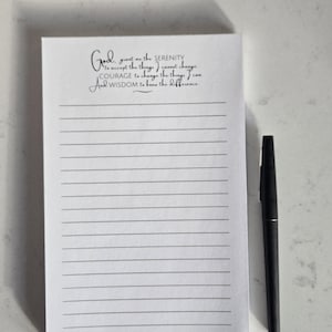 May include: A lined notepad with the text "God, grant me the serenity..." in elegant script. A black pen rests beside the notepad. The notepad is white with gray lines and text.