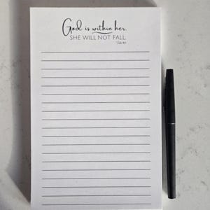 May include: A white lined notepad with the text "God is within her. SHE WILL NOT FALL" and a black pen. The notepad has a simple, elegant design, perfect for jotting down notes or journaling.