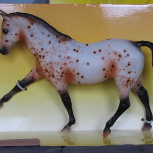 May include: A white and brown spotted horse figurine with black legs and a black mane and tail. The horse is standing on its hind legs with its front legs raised. The figurine is in a box with a yellow background.
