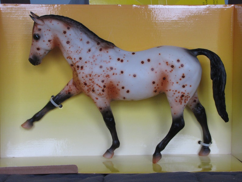 May include: A white and brown spotted horse figurine with black legs and a black mane and tail. The horse is standing on its hind legs with its front legs raised. The figurine is in a box with a yellow background.