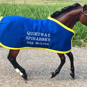 以下が含まれることがあります： 「Quietway Spinabbey Guy McLean」と刺繍された青と黄色の馬の毛布を着た茶色の馬のモデルです。