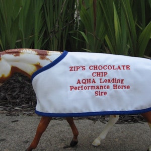 May include: A white horse blanket with blue trim and red embroidery that reads "Zip's Chocolate Chip AQHA Leading Performance Horse Sire".