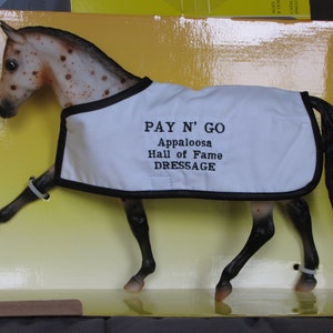 May include: A brown and white Appaloosa horse model wearing a white show blanket with black trim and the text "PAY N' GO Appaloosa Hall of Fame DRESSAGE".