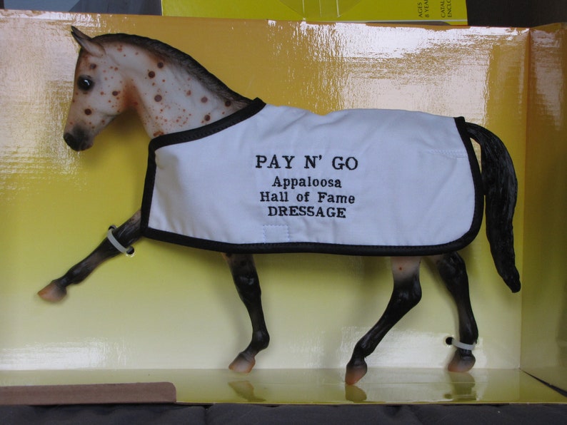 May include: A brown and white Appaloosa horse model wearing a white show blanket with black trim and the text "PAY N' GO Appaloosa Hall of Fame DRESSAGE".
