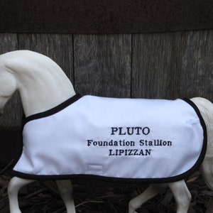 Puede incluir: Figura de caballo blanco con una manta blanca con ribete negro. La manta está bordada con el nombre "PLUTO", "Foundation Stallion" y "LIPPIZAN" en hilo negro. El caballo está posado sobre un fondo de madera.