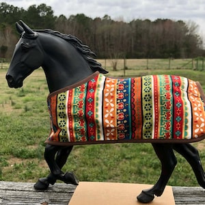 May include: A black horse figurine wearing a colorful patterned blanket. The blanket features vertical stripes in orange, red, green, and blue with floral and geometric designs. The horse is posed on a wooden surface with a green field and trees in the background.