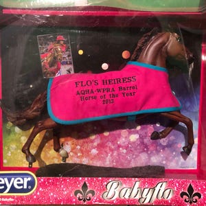 Könnte beinhalten: Ein Breyer-Modellpferd, "Flo's Heiress", in einer transparenten Kunststoff-Vitrine. Das Pferd ist braun mit einer rosa Decke mit blauer Bordüre. Auf der Decke steht "AQHA-WPRA Barrel Horse of the Year 2013."