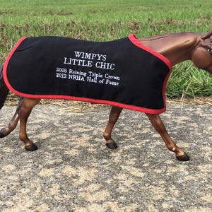 May include: A brown horse figurine wearing a black blanket with red trim. The blanket has white text that reads "Wimpys Little Chic" and lists "2008 Reining Triple Crown" and "2012 NRHA Hall of Fame."