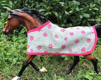 Op de afbeelding: Een bruin paard draagt ​​een wit en roze paardendek met een bloemenpatroon. Het deken heeft een roze rand en een witte afwerking.
