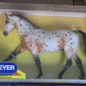 May include: A white and brown spotted Breyer model horse with a black mane and tail. The horse is standing on a yellow surface inside a clear plastic box. The box has a red, white, and blue label with the text "Breyer" and "No. 1116 Pay 'N Go Famous Dressage Appaloosa."