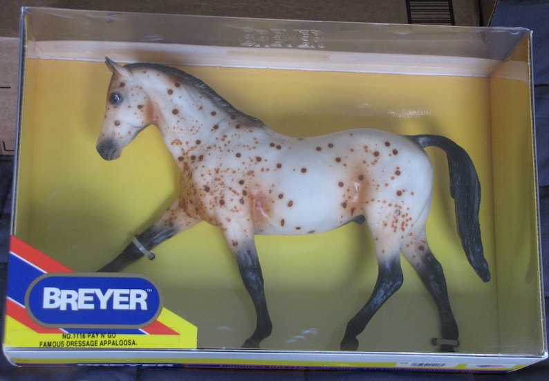 May include: A white and brown spotted Breyer model horse with a black mane and tail. The horse is standing on a yellow surface inside a clear plastic box. The box has a red, white, and blue label with the text "Breyer" and "No. 1116 Pay 'N Go Famous Dressage Appaloosa."
