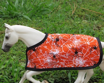 Op de afbeelding: Een oranje paardendeken met een wit spinnenwebontwerp en zwarte spinnen. De deken heeft een zwarte rand en wordt getoond op een wit paardenbeeldje. De achtergrond is groen gras.