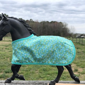 May include: A black horse figurine wearing a turquoise and floral patterned blanket. The blanket has a turquoise trim and is draped over the horse's back. The background features a cloudy sky and a blurred outdoor scene.