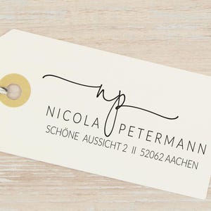 Könnte beinhalten: Ein rechteckiges, cremefarbenes Geschenkanhänger-Etikett aus Papier mit einer goldfarbenen, verstärkten Öse und einer schwarz-weißen Schnur. Das Etikett trägt den schwarzen Aufdruck NICOLA PETERMANN, die Adresse SCHÖNE AUSSICHT 2, 52062 AACHEN und ein geschwungenes np-Monogramm.