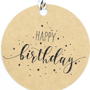 Könnte beinhalten: Ein runder, brauner Papieretikett mit einer schwarzen Schnur. Das Etikett trägt den Text "HAPPY birthday" in schwarzer Kursivschrift mit schwarzen Punkten, die um den Text verstreut sind.