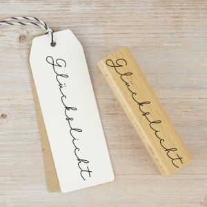May include: A wooden stamp with the text "Gl&uuml;cklich" in cursive script. The stamp is on a light wood surface.