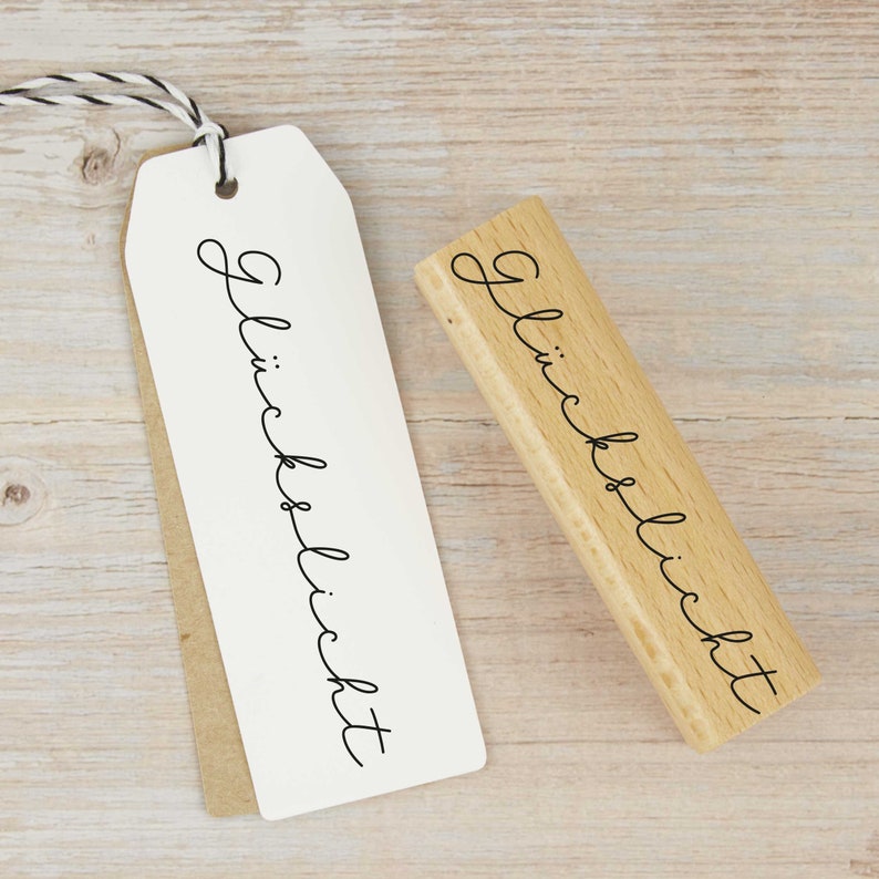 May include: A wooden stamp with the text "Gl&uuml;cklich" in cursive script. The stamp is on a light wood surface.