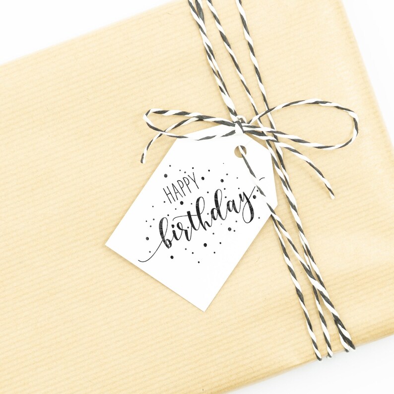 May include: A brown paper gift bag with a white tag tied with black and white twine. The tag has the text "Happy birthday" written in black cursive with black dots scattered around the text.