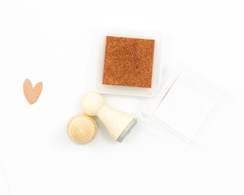 以下が含まれることがあります： 茶色のインクパッドが付いた木製スタンプのセット。スタンプはハート型と円形です。インクパッドは透明なプラスチックケースに入っています。
