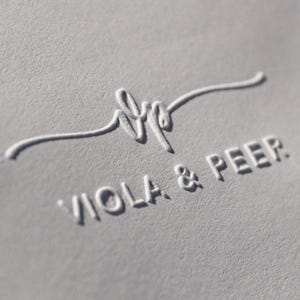 Könnte beinhalten: Eine hellgraue, texturierte Papieroberfläche mit einem geprägten Logo, das ein stilisiertes Schreibschrift-Monogramm über dem Text VIOLA & PEER zeigt. Das Design erzeugt einen subtilen dreidimensionalen Effekt durch Schattenwurf.