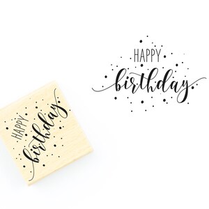 May include: A wooden stamp with the text "Happy birthday" in a cursive font with black ink and small black dots scattered around the text.