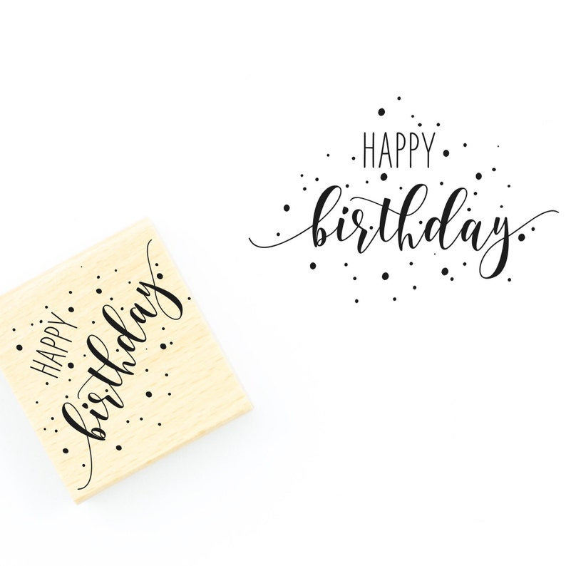 May include: A wooden stamp with the text "Happy birthday" in a cursive font with black ink and small black dots scattered around the text.