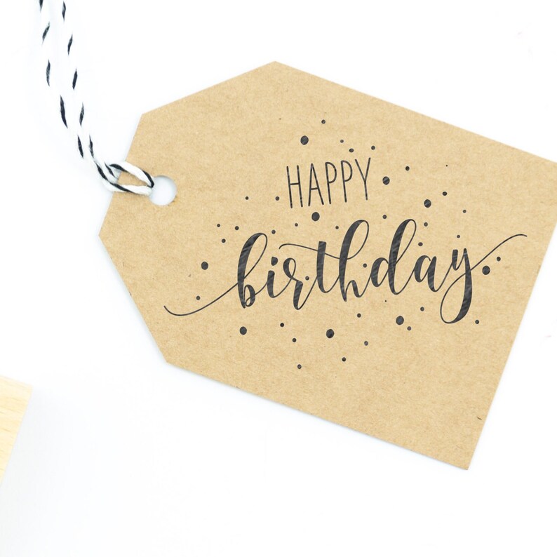 May include: A brown paper gift tag with a white string attached. The tag has the text "Happy birthday" written in black cursive with black dots scattered around the text.