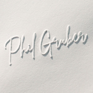 Könnte beinhalten: Nahaufnahme eines weißen Papiers mit der geprägten Unterschrift "Phil Gerber" in Schreibschrift. Die Signatur befindet sich in der Mitte, die Papierstruktur ist sichtbar.