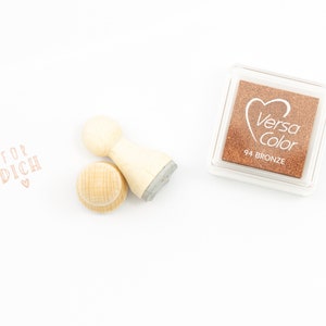 以下が含まれることがあります： 「FOR DICH」という文字と小さなハートが刻まれた木製スタンプ。スタンプの隣には「Versa Color 94 BRONZE」と書かれた茶色のインクパッドがあります。