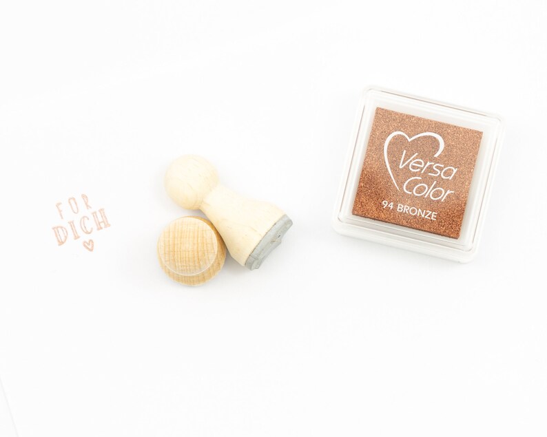以下が含まれることがあります： 「FOR DICH」という文字と小さなハートが刻まれた木製スタンプ。スタンプの隣には「Versa Color 94 BRONZE」と書かれた茶色のインクパッドがあります。