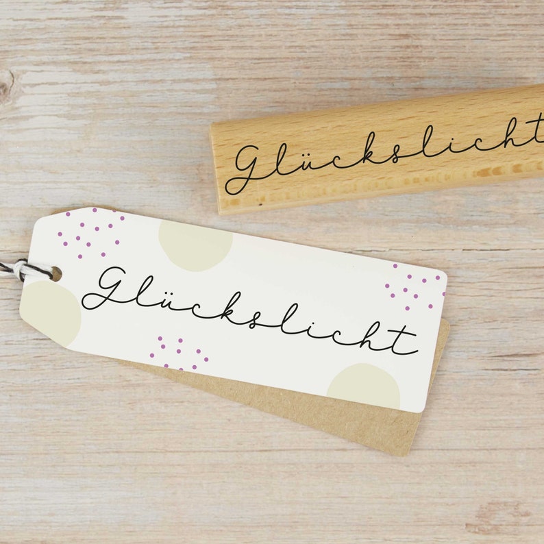 May include: A wooden stamp with the word "Gl&uuml;cklich" written in cursive. The stamp is on a light wood surface with a white card with the same text and a purple polka dot pattern.