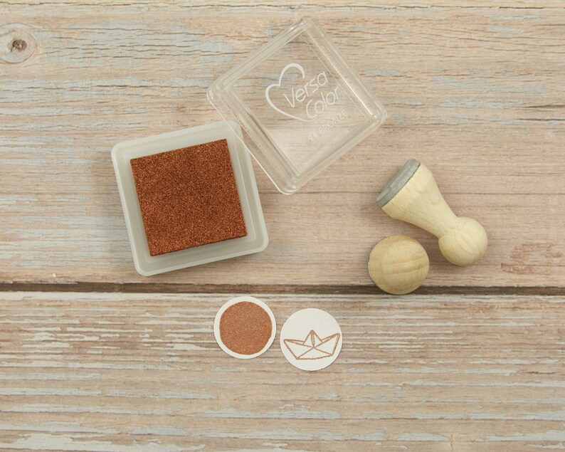 以下が含まれることがあります： スタンプ用のクラフト用品セット。茶色のインクパッド、紙のボートのデザインが施された木製スタンプ、木製スタンプハンドルが含まれています。インクパッドは、透明なプラスチックケースに入っていて、ケースには「Versa Color」の文字が書かれています。
